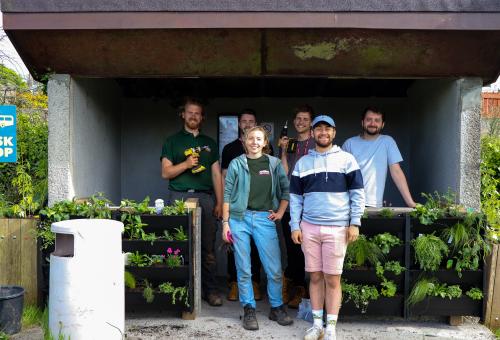 6 Eden apprentices smile in front of their newly transformed "Buzz Stop". A local bus stop can be seen with plants in planters on its walls.